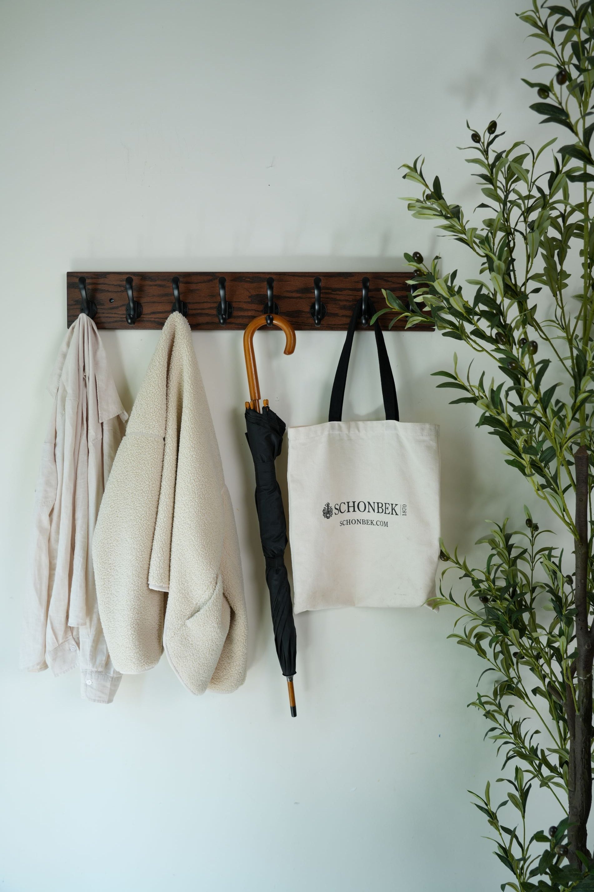 Wooden coat rack with folded clothes and an umbrella against a white wall with greenery at the bottom.