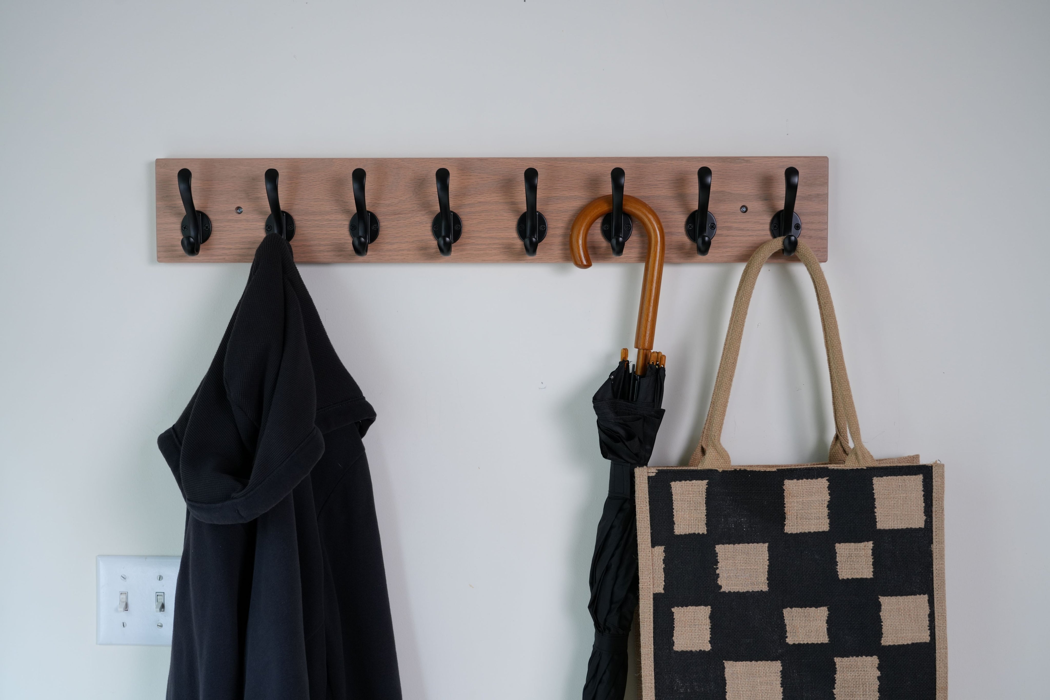 Wooden coat rack with hooks holding a black coat, brown umbrella, and checkered bag against a white wall.